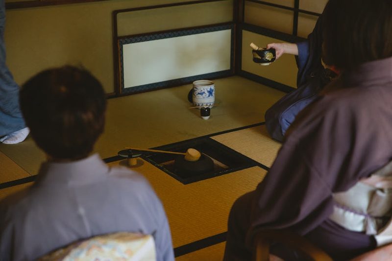 Anonymous group of people wearing authentic robe sitting near bamboo ladle on iron pot and dishware for traditional Japanese ceremony