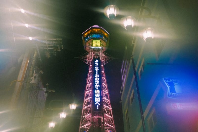 A vibrant night view of Tsutenkaku Tower in Osaka, Japan, illuminated with colorful neon lights.