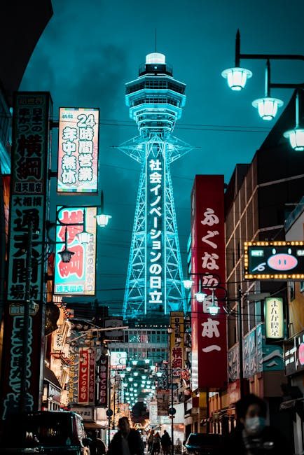 Illuminated Tsūtenkaku Tower amidst bustling urban street in Osaka at night.