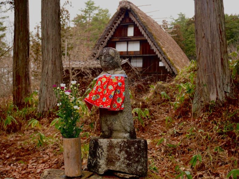 Stone statue wearing traditional clothing in Takayama garden with flowers and thatched roof house.