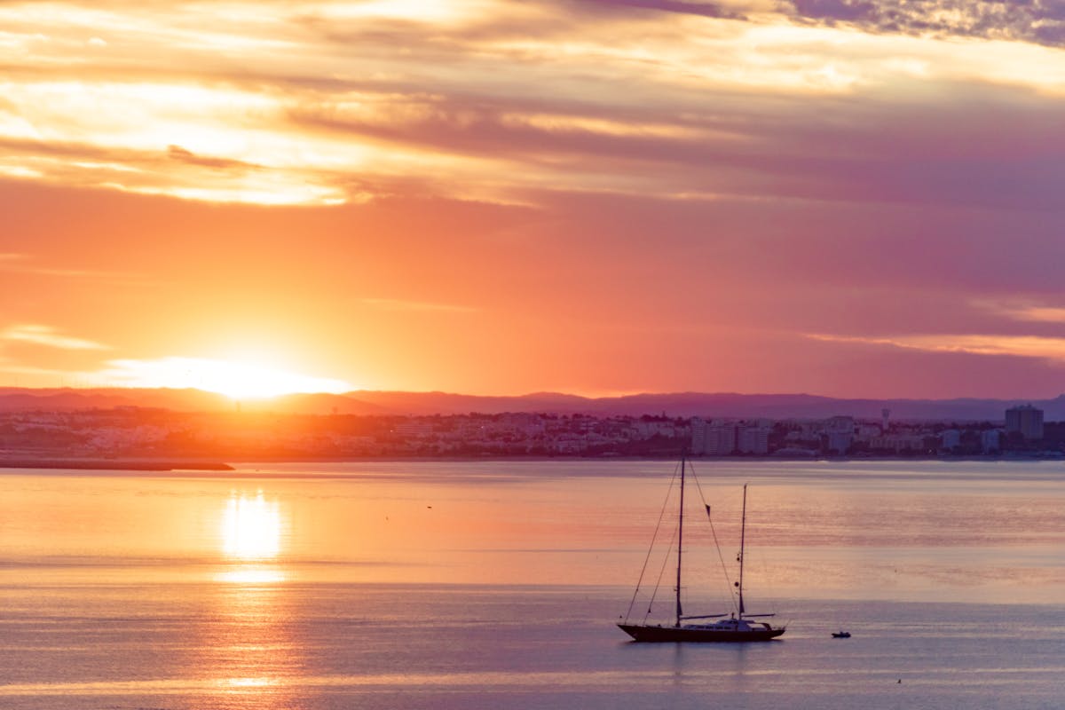 A serene sunset over the Algarve coast near Lagos with a silhouetted sailboat on calm water