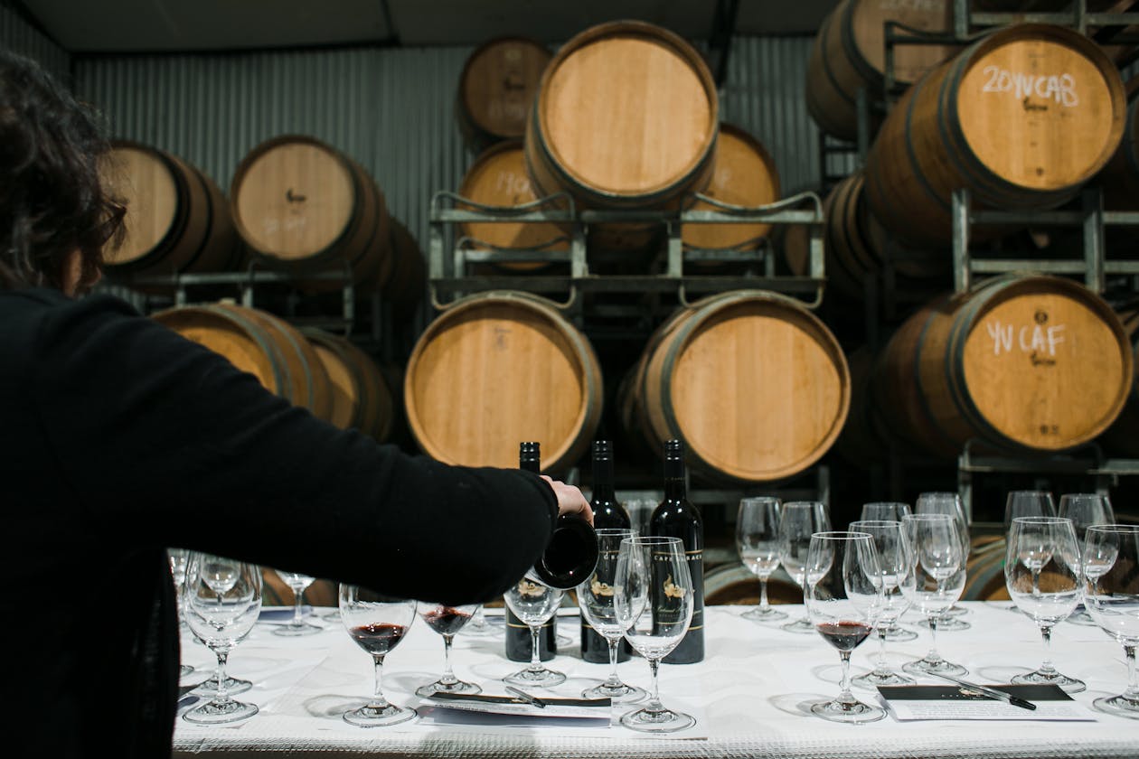 A sommelier pouring wine from a bottle into glasses with wooden barrels visible in the background