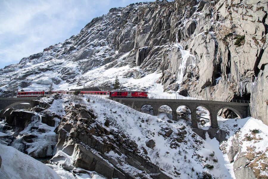 Red train crossing a dramatic stone viaduct in snowy Alpine mountains entering a tunnel