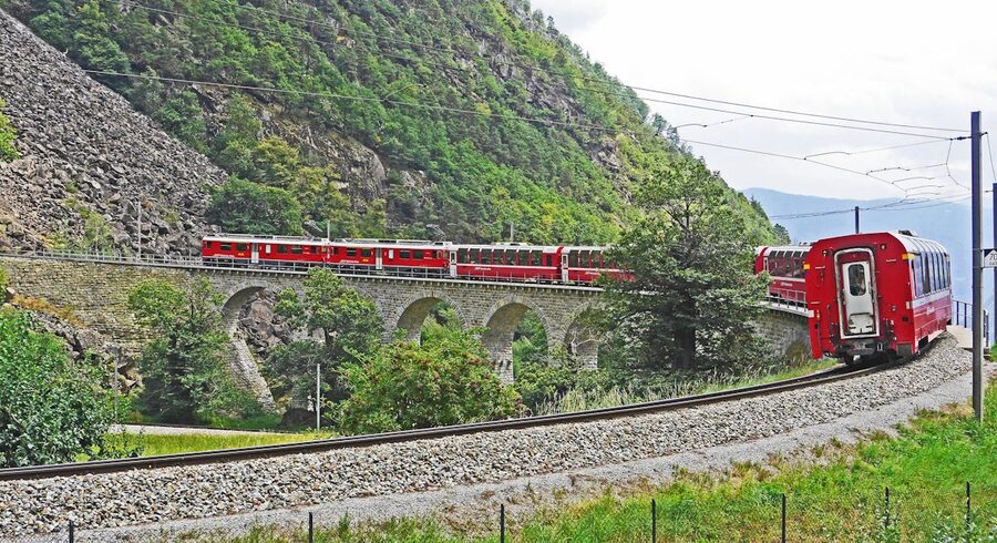 Red train crossing a sweeping stone viaduct in a lush green mountain valley