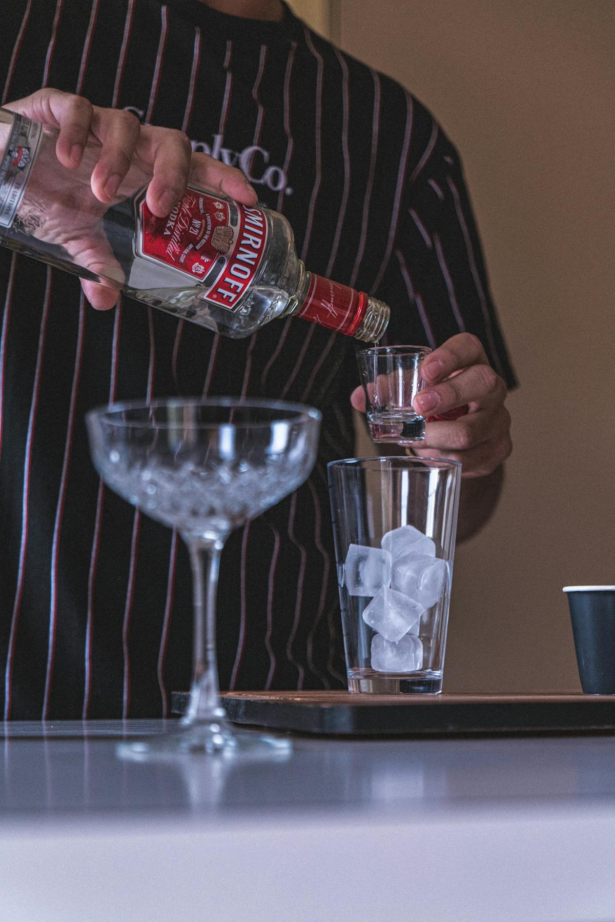 Bartender pouring vodka into a glass with ice cubes at a bar