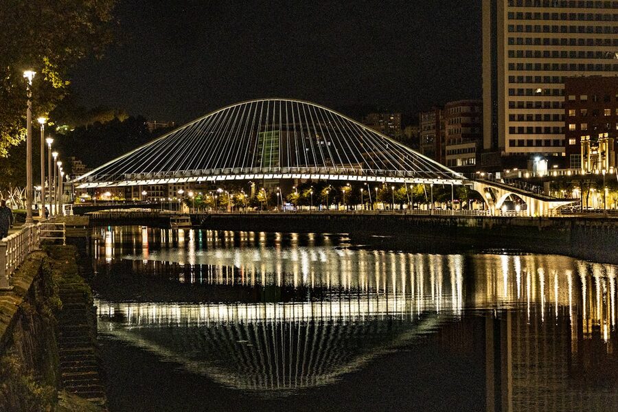 The Zubizuri Bridge in Bilbao lit up at night with reflections shimmering in the Nervion river below