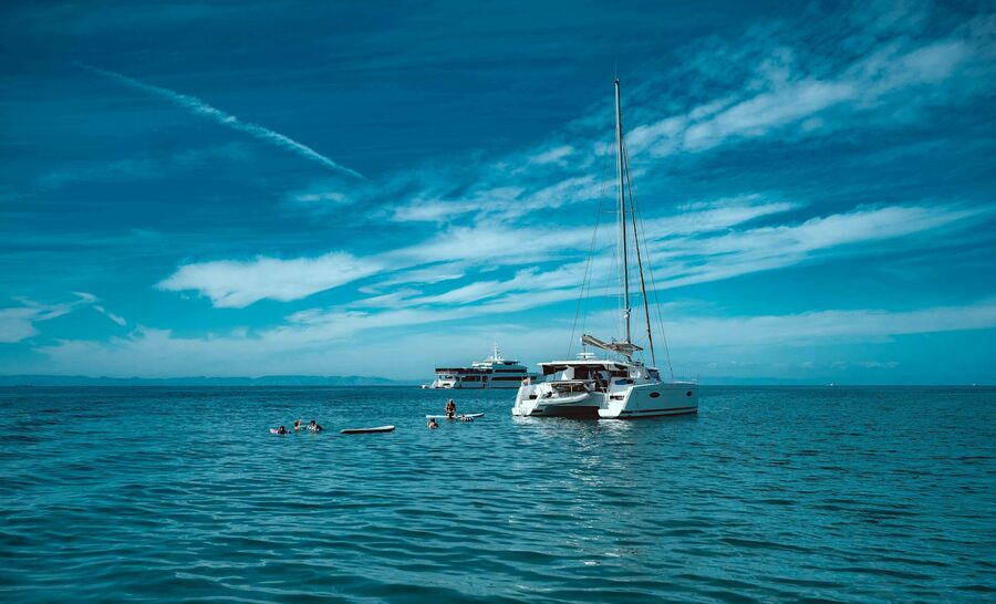 Catamaran sailing in open sea with people swimming nearby under clear blue sky