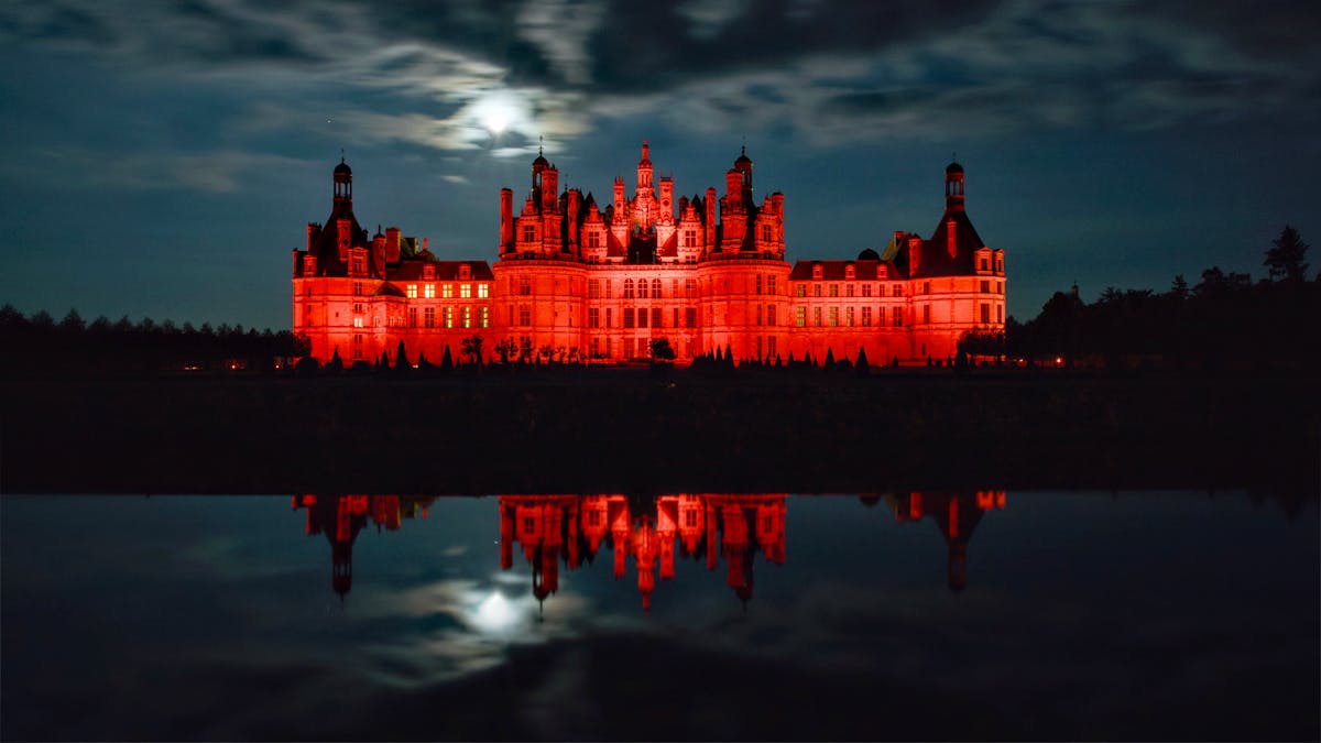 Chambord castle glowing red under night sky with reflection in water