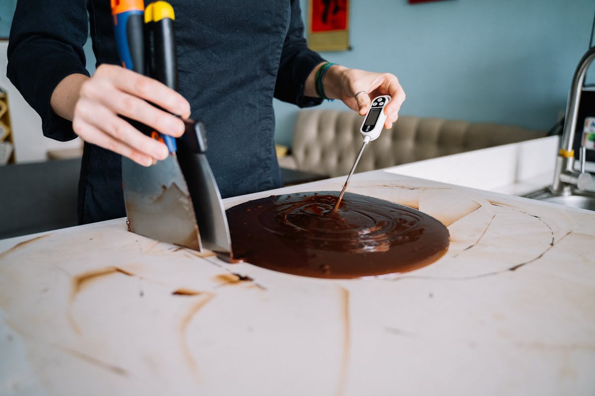 Person tempering chocolate on a kitchen counter using a scraper and thermometer
