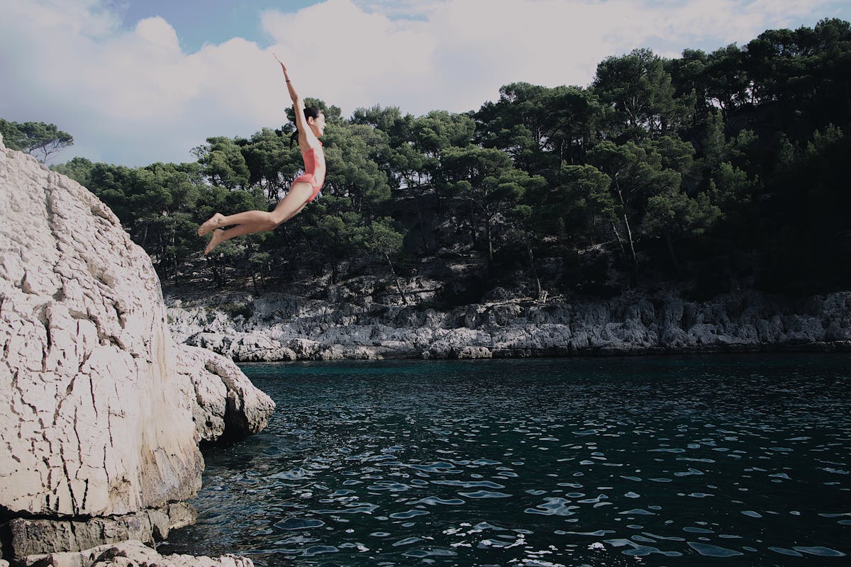 Woman in swimsuit diving from rocky cliff into the sea surrounded by greenery