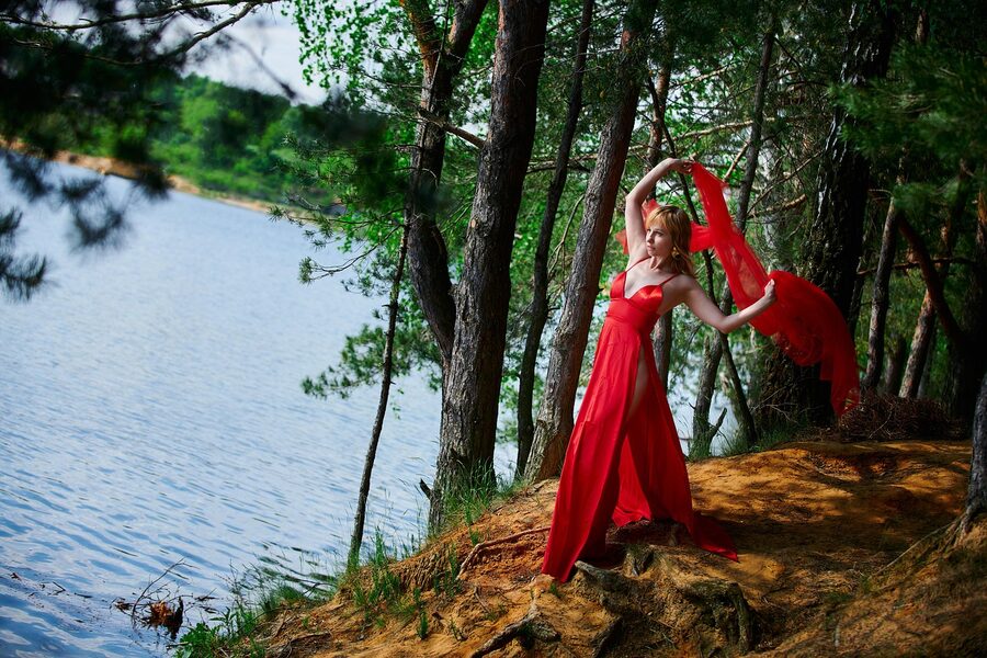A woman in a flowing red flamenco dress dancing by a lake surrounded by trees