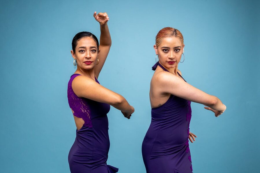 Two women performing flamenco in purple costumes against a blue background