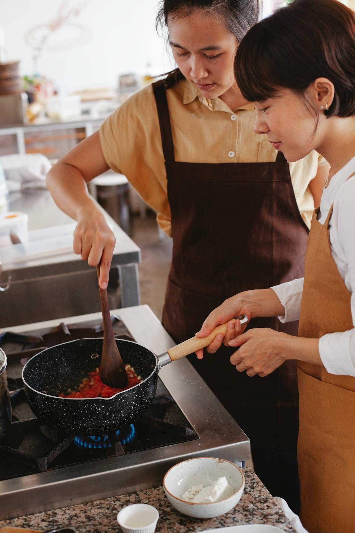 Two women cooking together during a culinary class wearing aprons in a kitchen