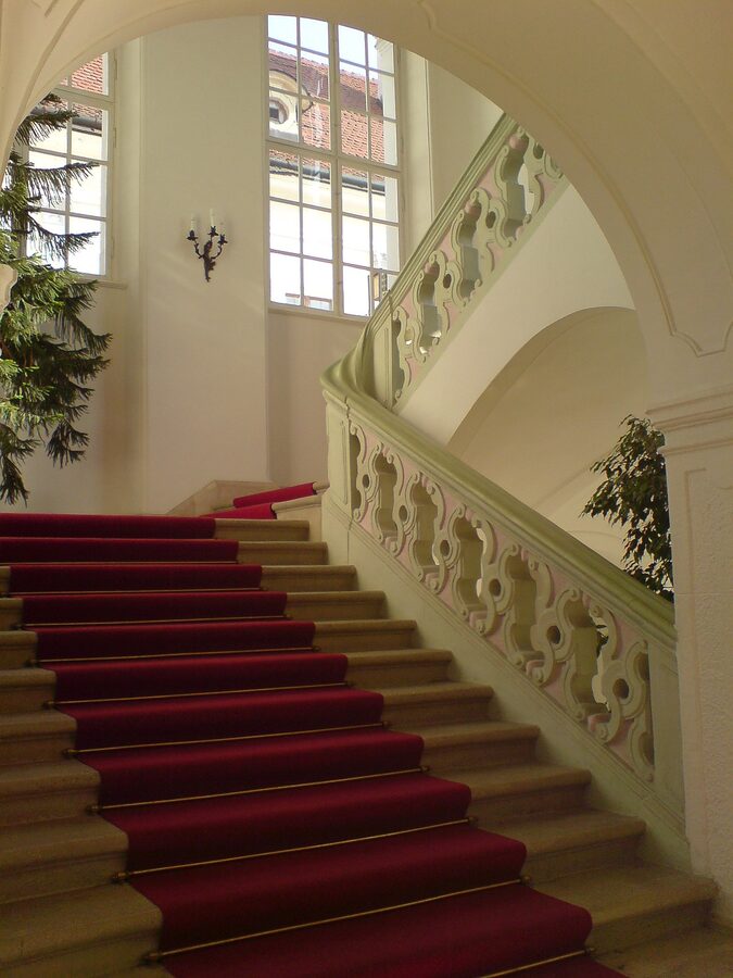 Grand staircase with red carpet and ornate balustrade inside Gödöllő Palace