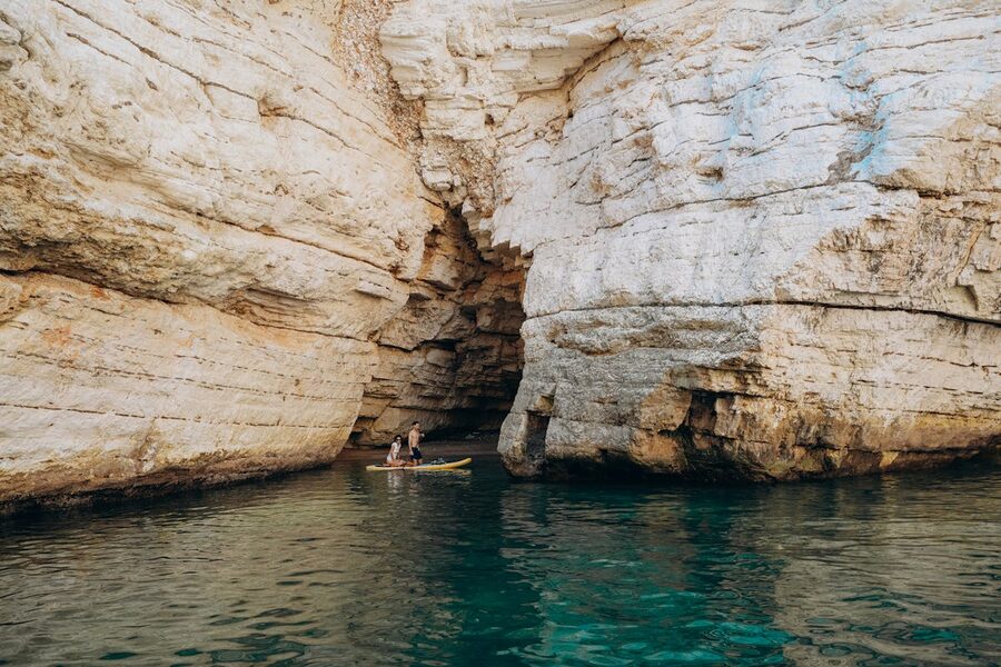 Group of kayakers paddling into a large limestone cave opening on a Mediterranean coast