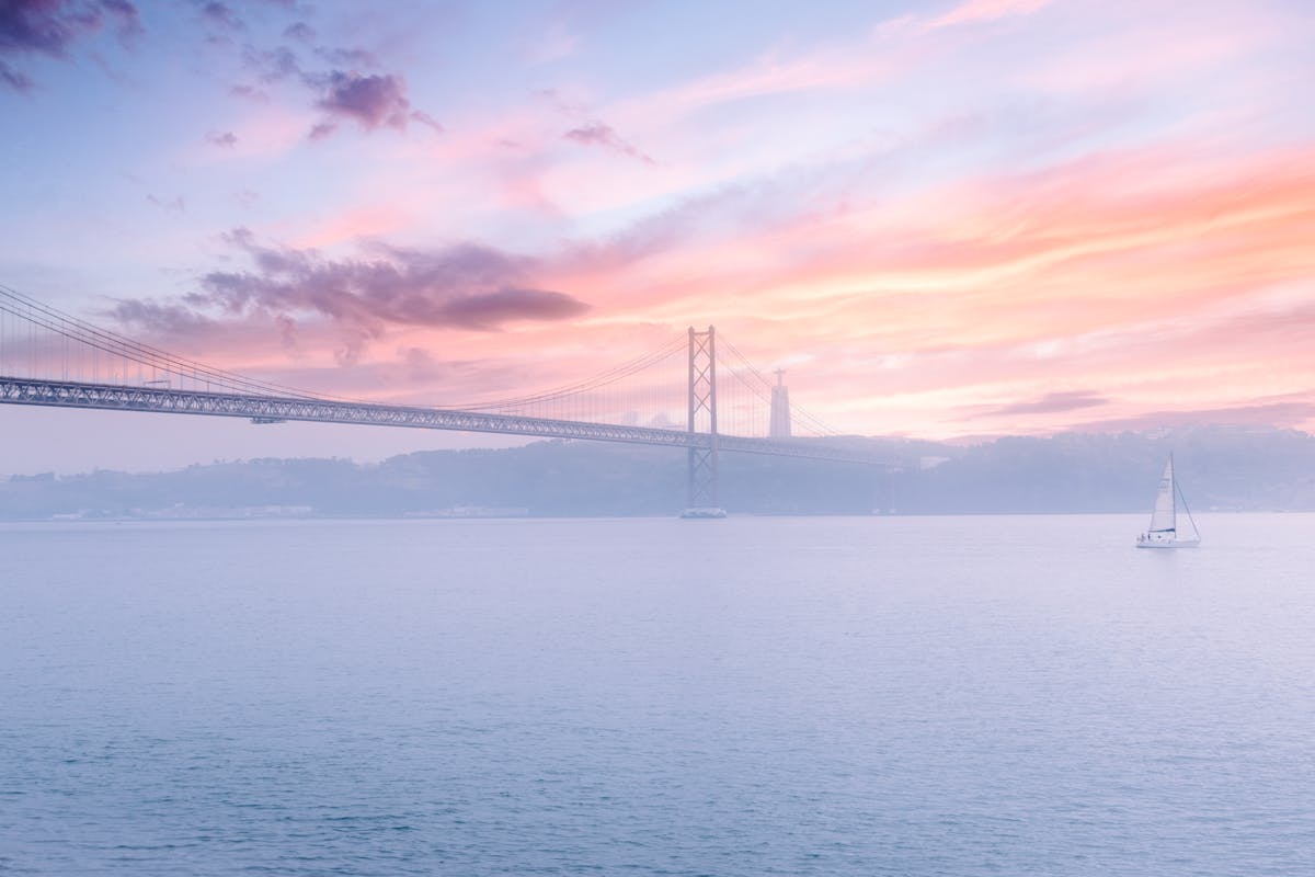 A sailboat on calm waters of the Tagus River with the 25 de Abril Bridge at sunset