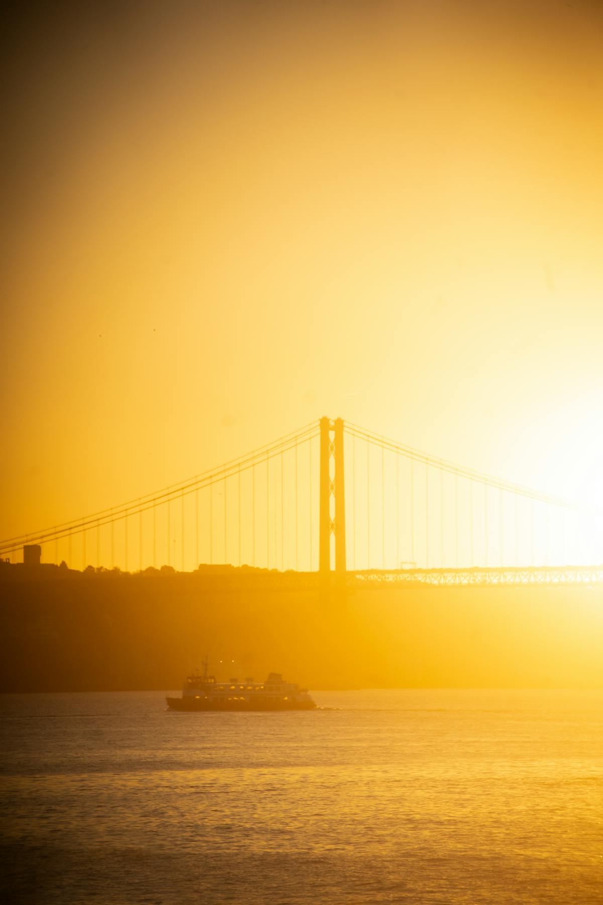 Silhouette of a ferry passing under the 25 de Abril Bridge in Lisbon at sunset