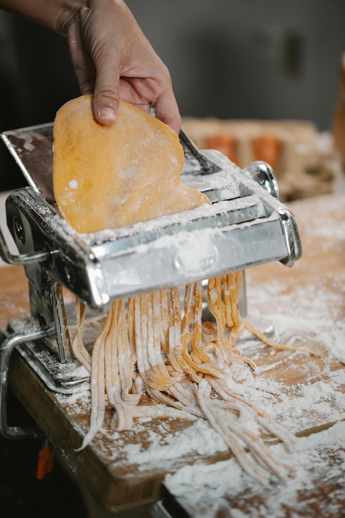 Person cutting fresh pasta dough with a pasta machine on a wooden table covered in flour