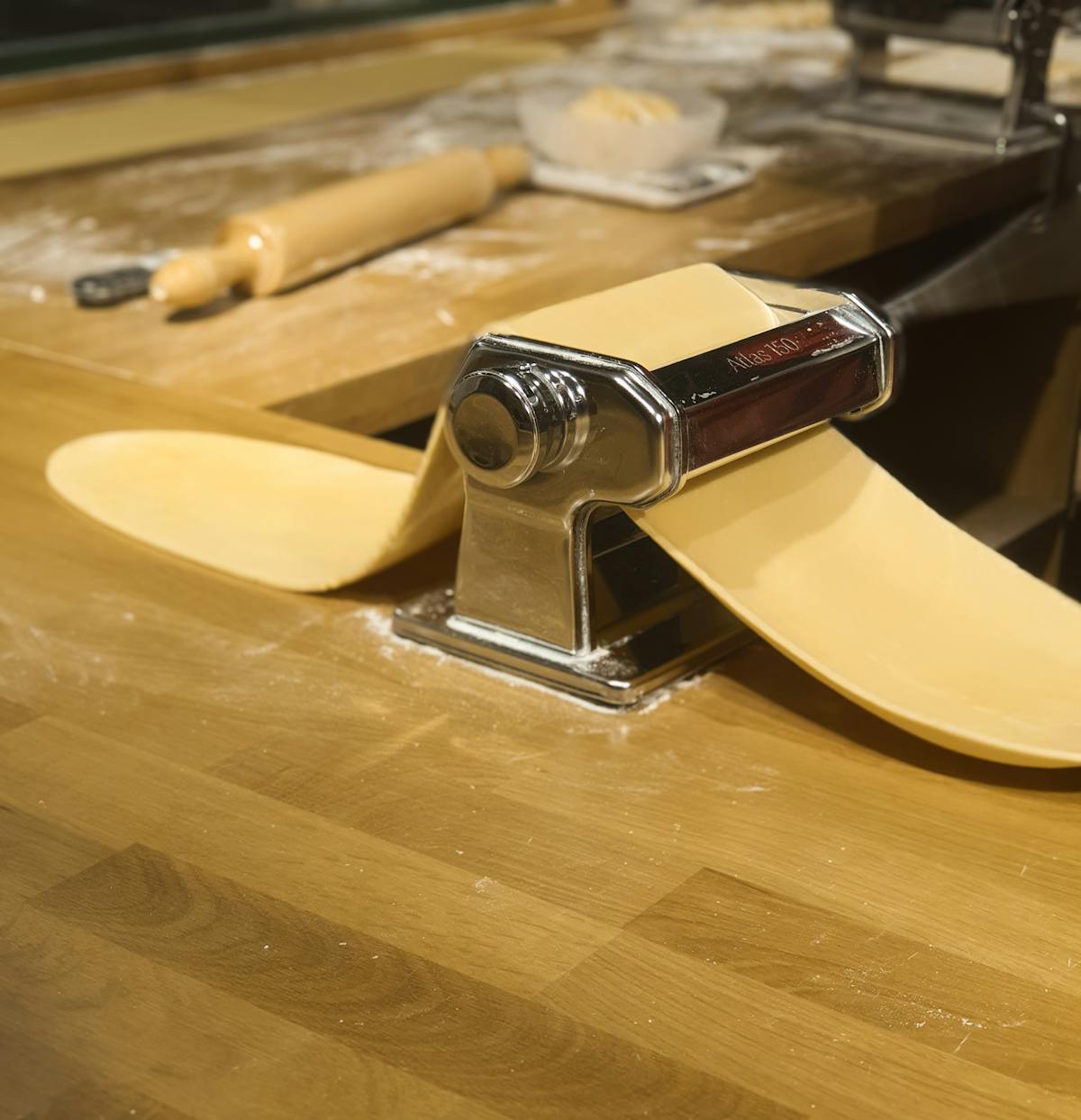 Fresh pasta emerging from a hand-cranked pasta machine on a flour-dusted wooden countertop