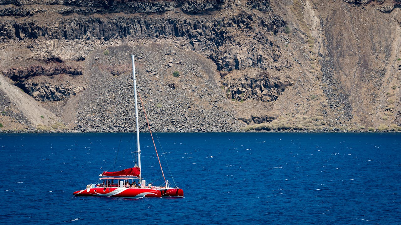 Red sailboat in deep blue Santorini waters with rocky cliffs behind