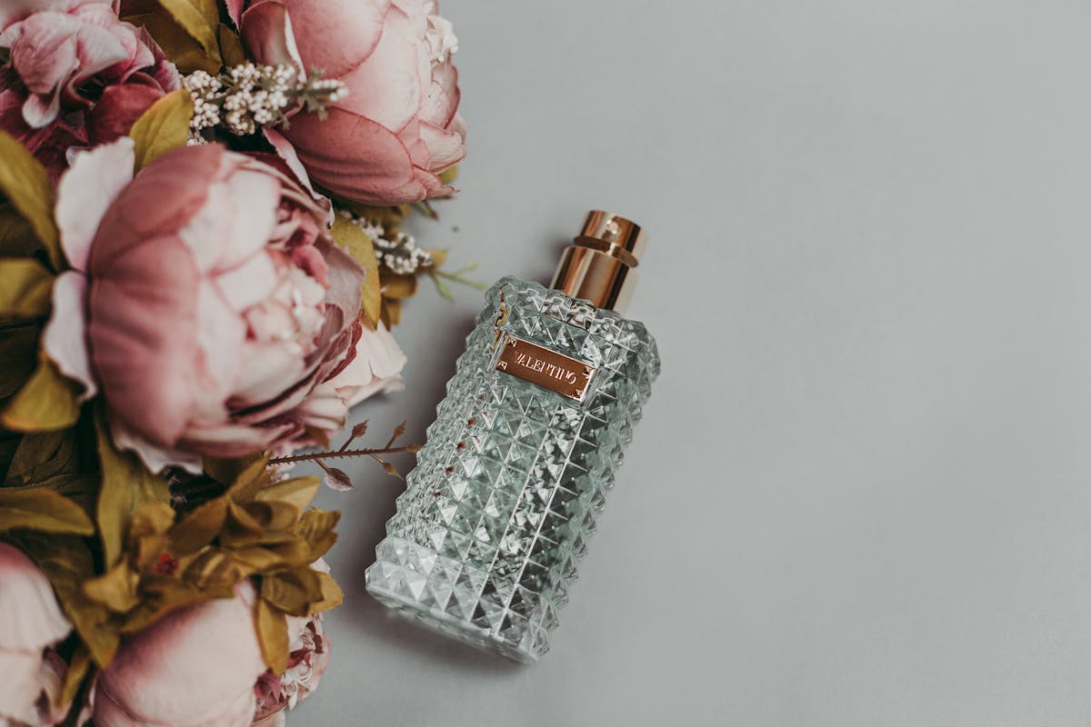 Glass perfume bottle beside pink peony flowers on grey background