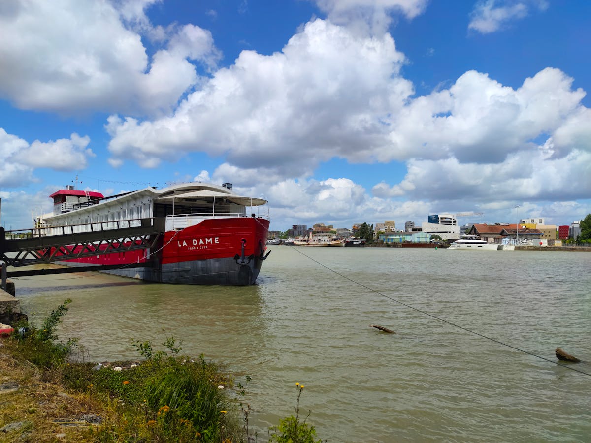 A red cruise boat named La Dame docked on the Garonne River in Bordeaux with city buildings behind