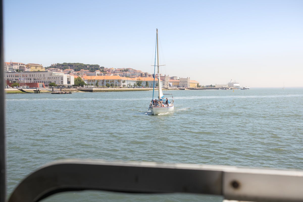 A sailboat carrying travelers navigates the Lisbon waterfront under a clear sky