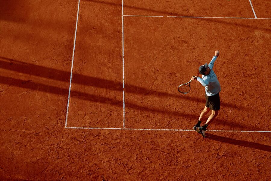 Tennis player serving on a red clay court with long shadows at sunset
