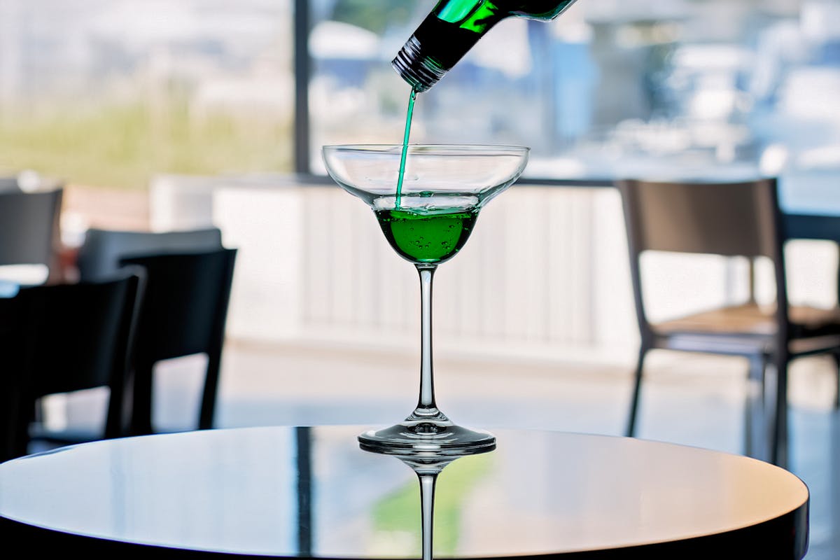 A green cocktail being poured into a glass with a blurred bar background