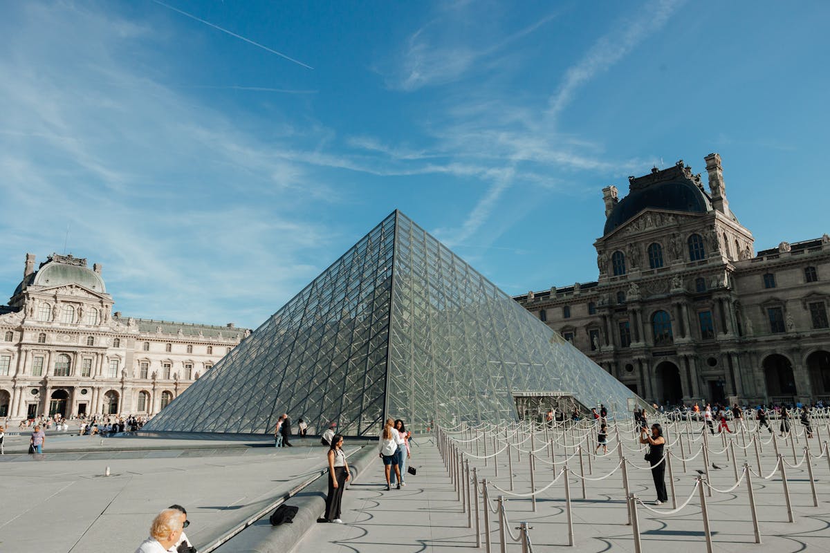 Visitors walking near the Louvre Pyramid on a sunny day with blue sky and historic buildings
