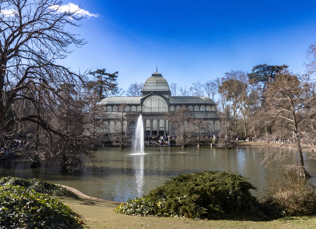 The Crystal Palace in Retiro Park Madrid with a tranquil pond and fountain under a clear sky