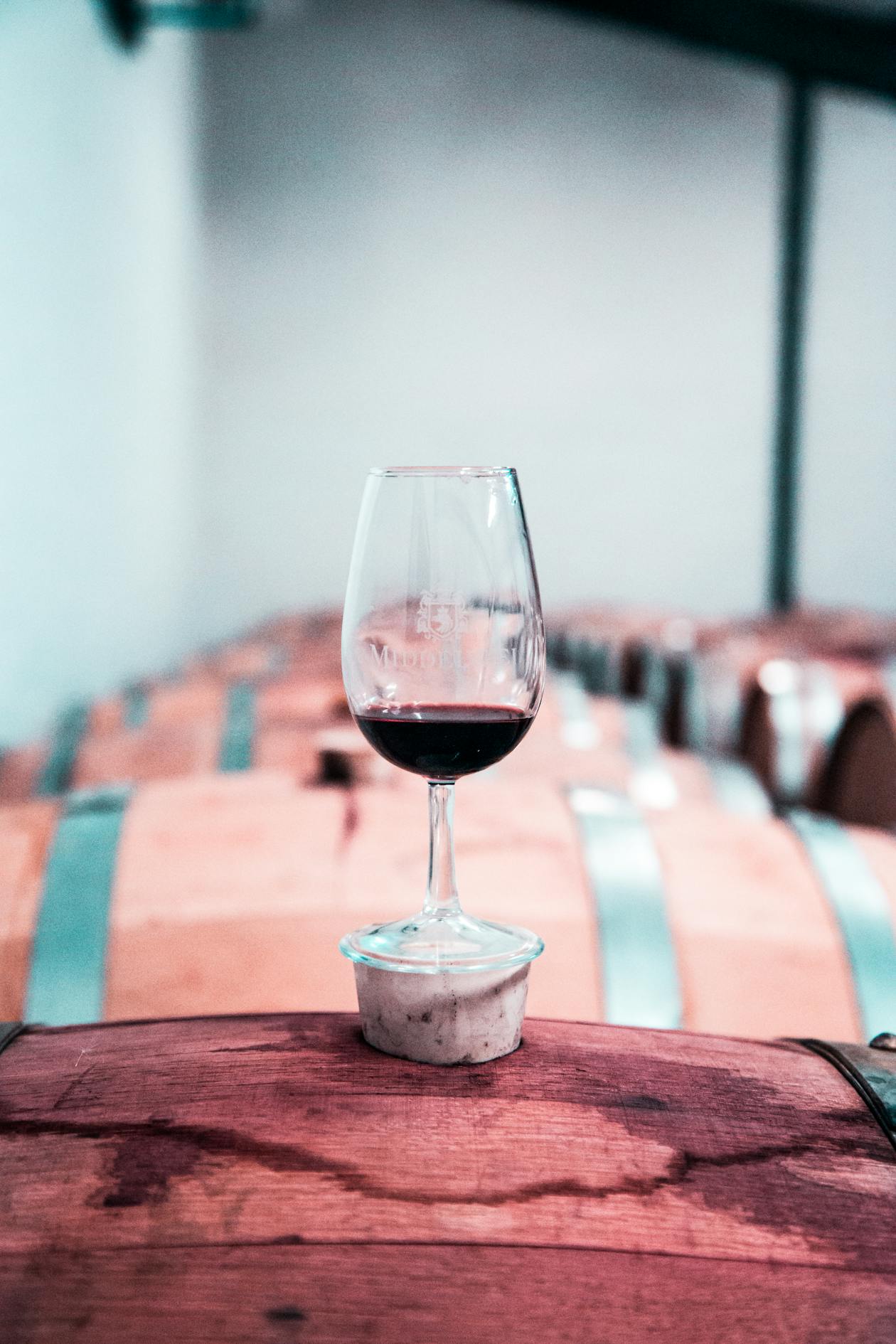 A single glass of red wine resting on an oak barrel in a dimly lit cellar