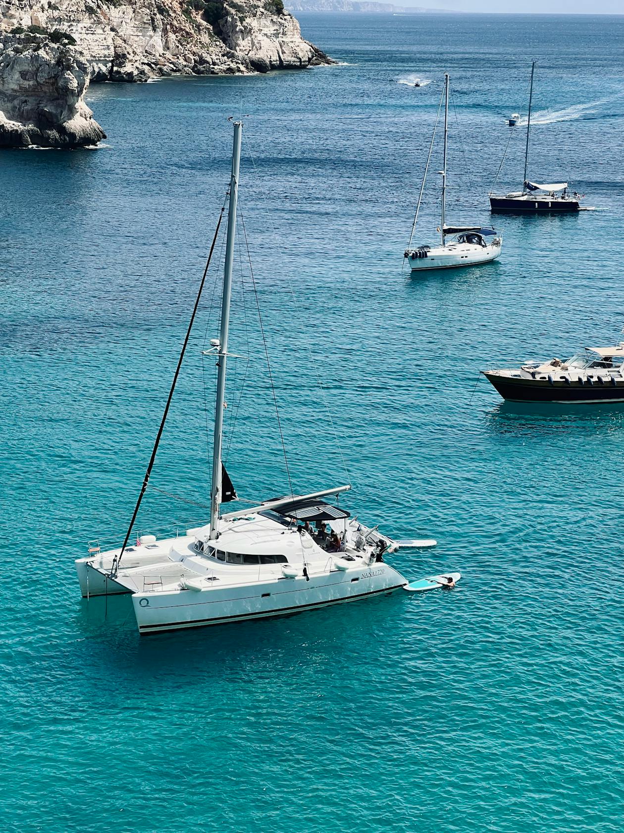 Yacht and sailboats cruising through clear turquoise water near rocky coastline