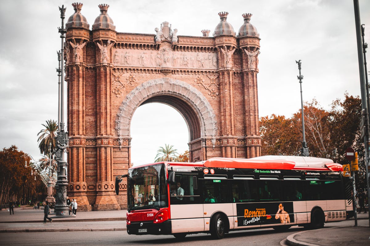 Red city bus passing by the historic Arc de Triomf in Barcelona with autumn leaves