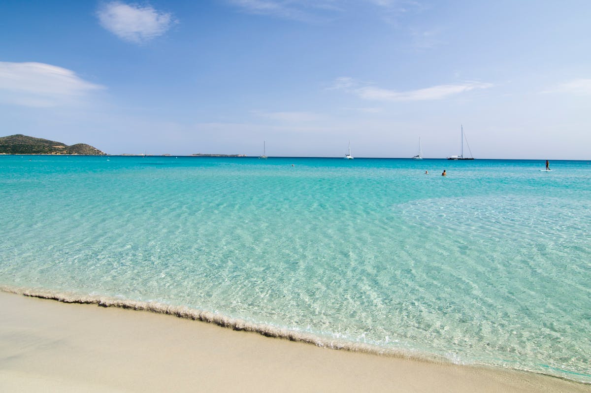Pristine turquoise beach with sailboats in the distance off the Sardinian coast
