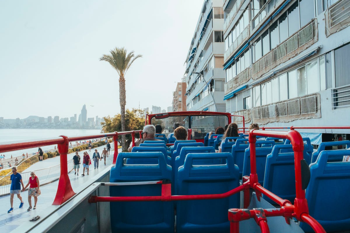 Tourists on an open-top double-decker bus tour with a beachfront view on a sunny day