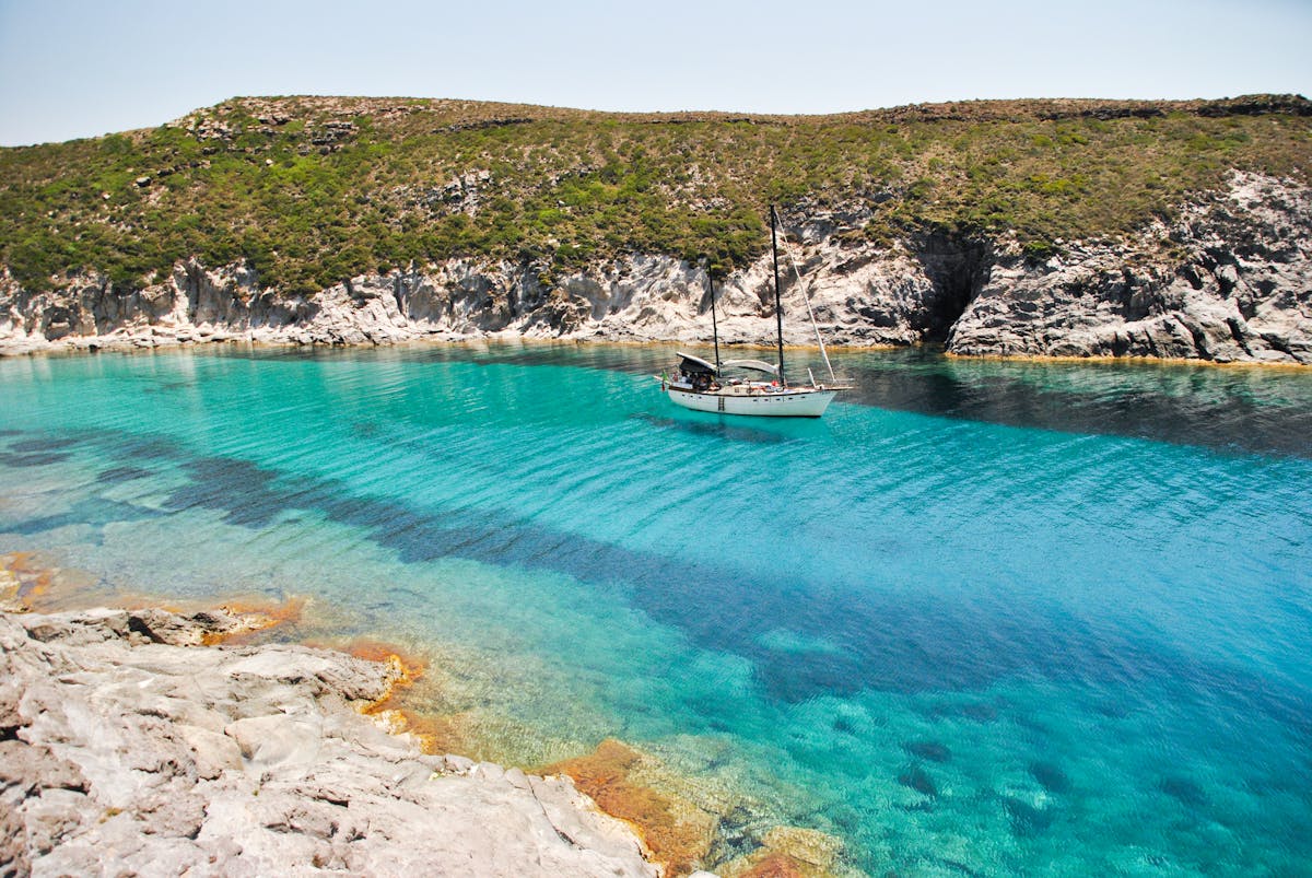 Sailboat floating on crystal clear blue waters near the rocky Sardinian coastline