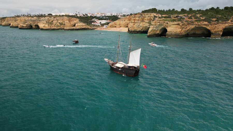 A vintage pirate ship sails past dramatic rocky cliffs along the Algarve coastline