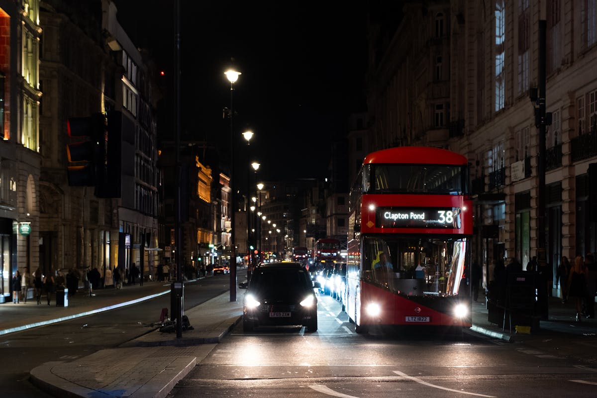 A red double-decker bus on a busy London street at night with illuminated buildings