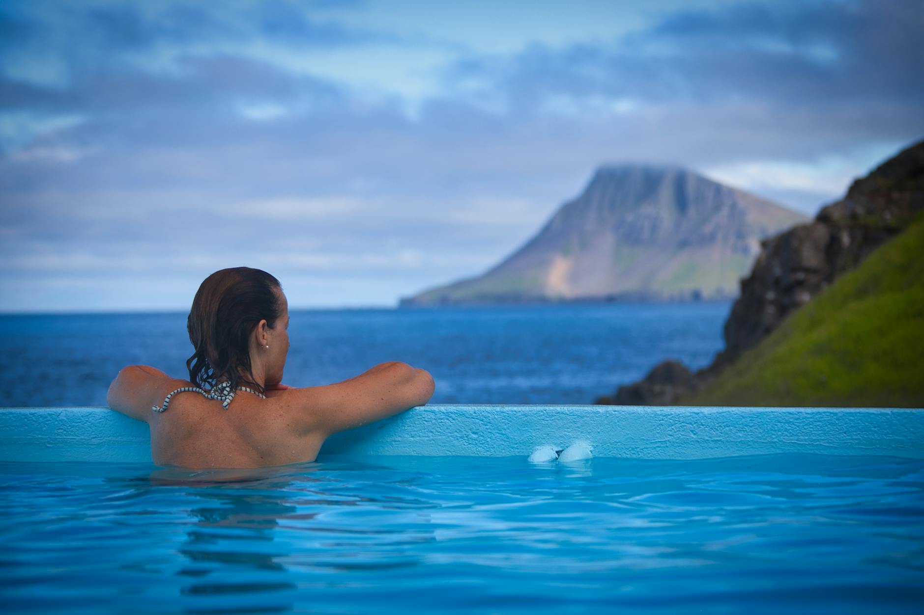 Back view of a woman relaxing in outdoor thermal pool with Icelandic mountain scenery