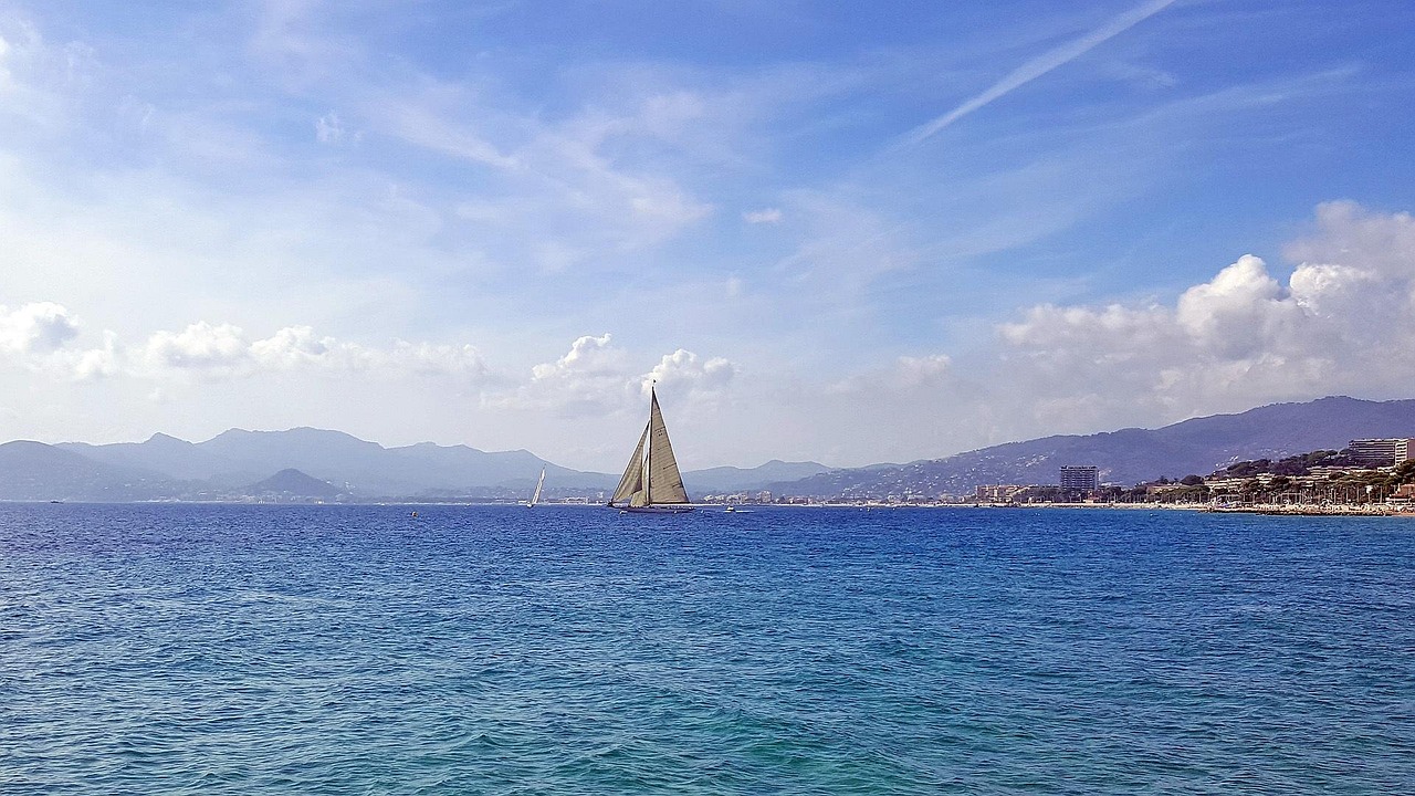 White sailboat on calm blue water with the Cannes shoreline visible behind