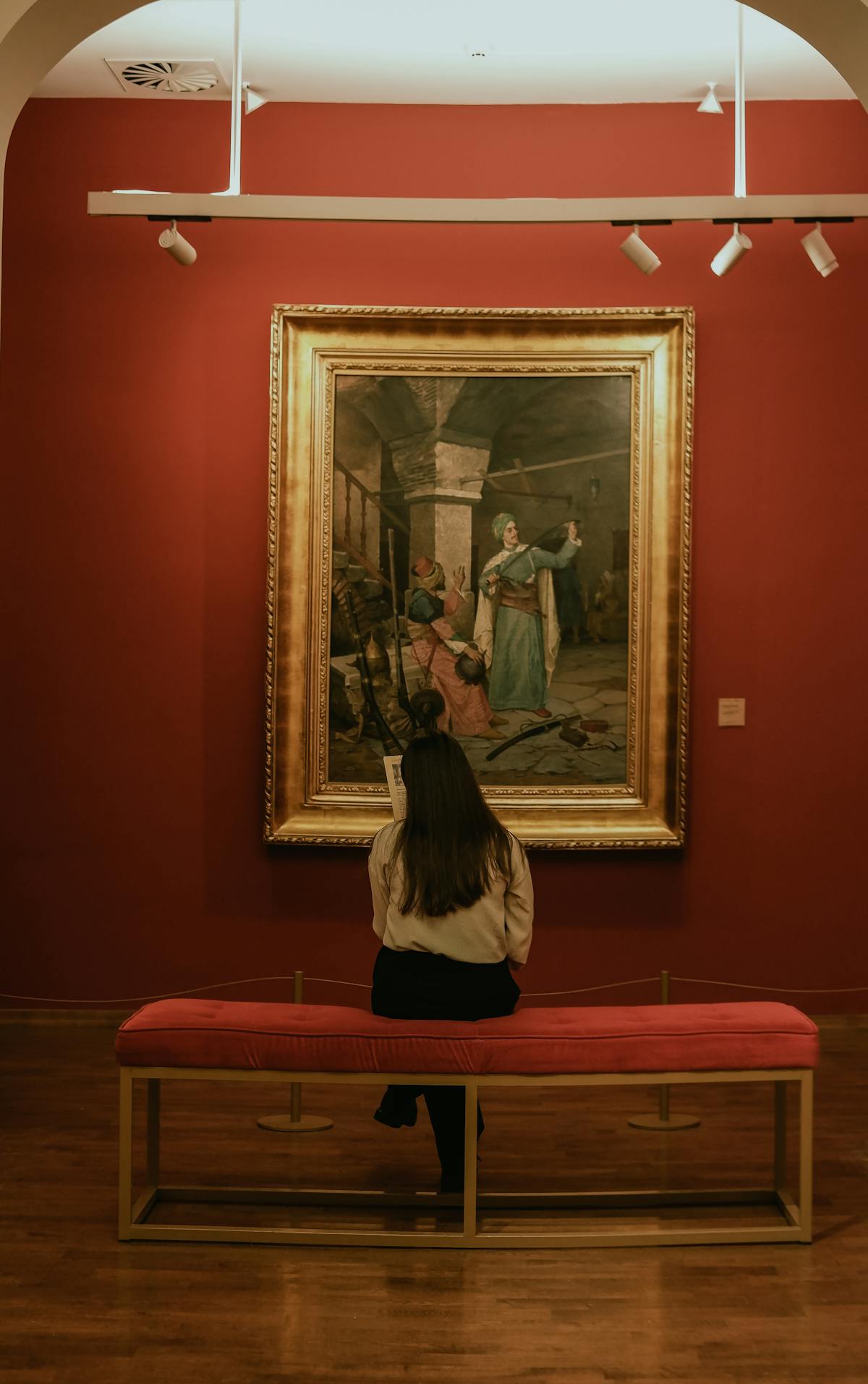 A woman observes a classical painting in a museum gallery with red walls