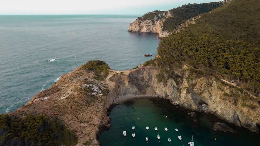 Aerial view of the sheltered Sa Tuna bay with boats anchored in turquoise water along the Costa Brava coastline