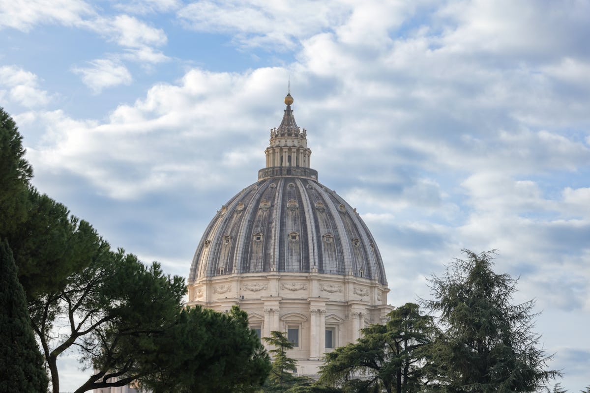 The exterior of St. Peters Basilica dome surrounded by trees against a blue sky