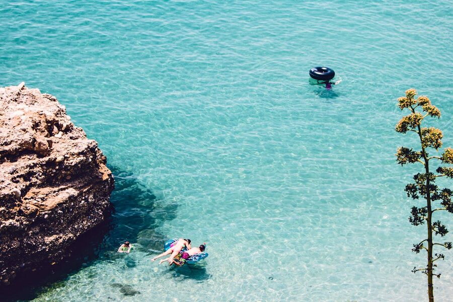 People swimming and floating in crystal clear blue water beside a rocky Mediterranean coastline
