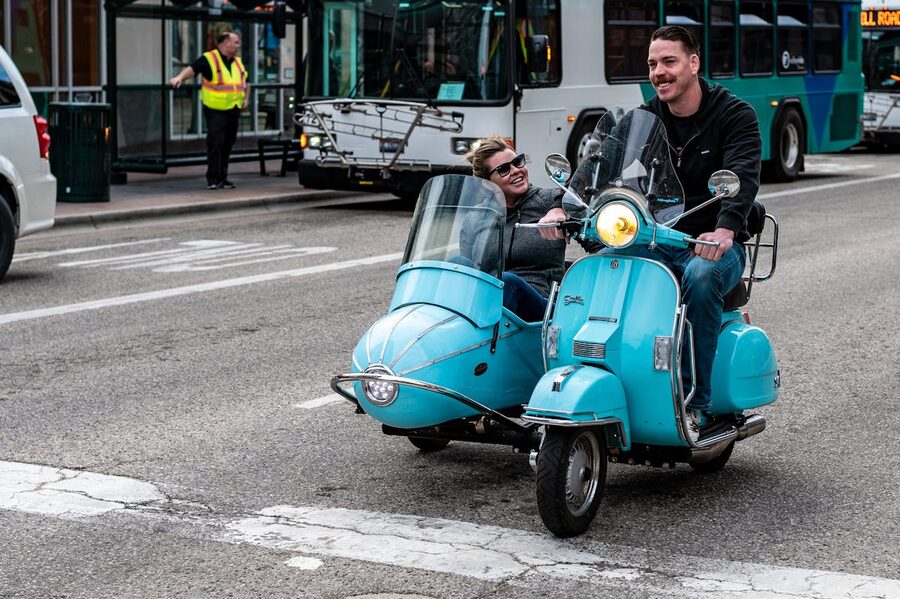 Joyful couple riding a blue vintage scooter with sidecar through city streets