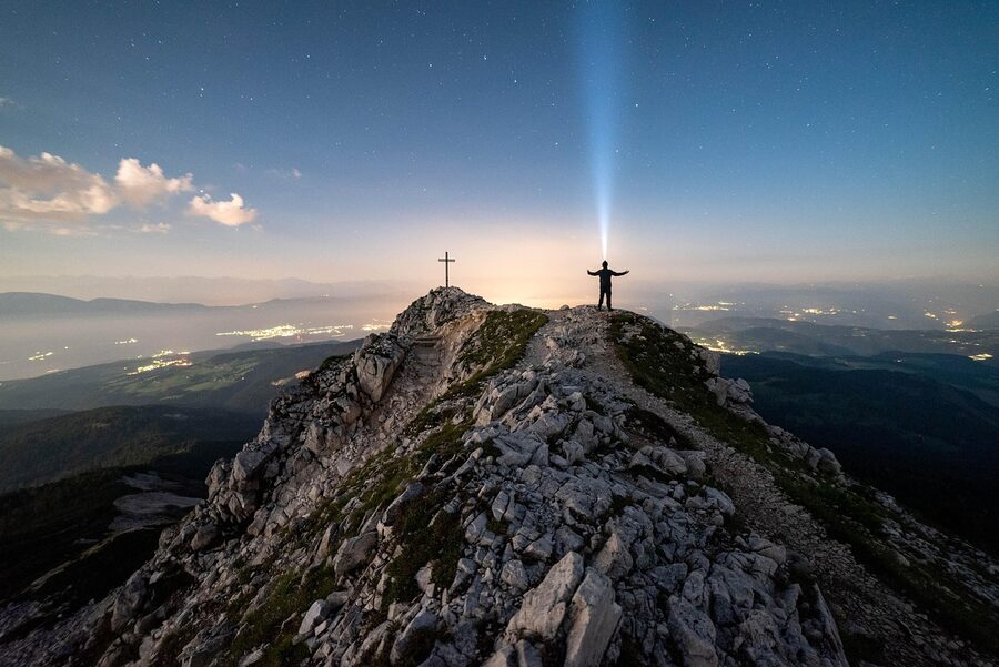 A man with a headlamp at a mountain summit under the Milky Way and stars